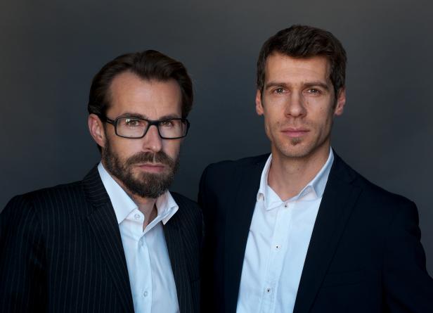 Zwei Männer in Anzügen stehen vor einem grauen Hintergrund.