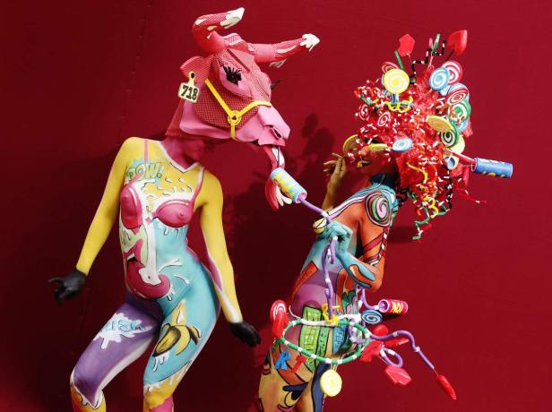 Zwei Frauen mit Körperbemalung und extravaganten Kopfbedeckungen posieren vor einem roten Hintergrund.