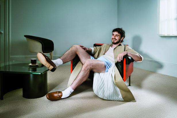 Ein Mann sitzt lächelnd in einem Sessel, bekleidet mit Shorts, Socken und einem Trenchcoat.