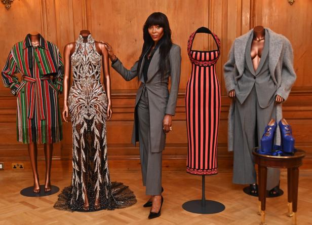 Naomi Campbell posiert neben mehreren Mannequins mit verschiedenen Kleidungsstücken.