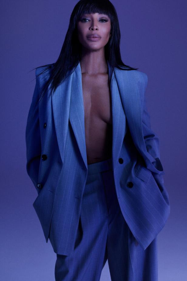 Naomi Campbell posiert in einem blauen Hosenanzug vor einem violetten Hintergrund.