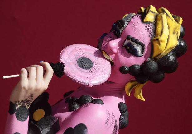 Eine Person mit rosa Körperbemalung und einem übergroßen Lolli vor rotem Hintergrund.