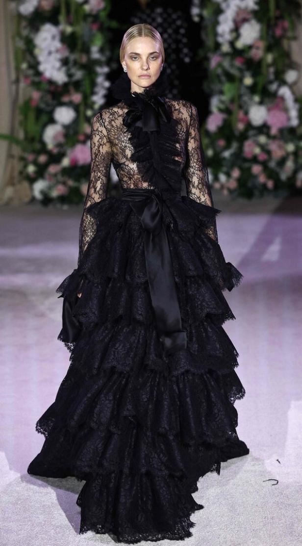 Ein Model präsentiert ein schwarzes, bodenlanges Spitzenkleid mit Rüschen und Schleife auf dem Laufsteg.