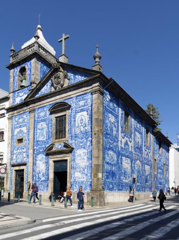 Igreja Santo Ildefonso mit blauer Fassade,bei Sonnenschein. Davor eine Straße auf der Menschen gehen