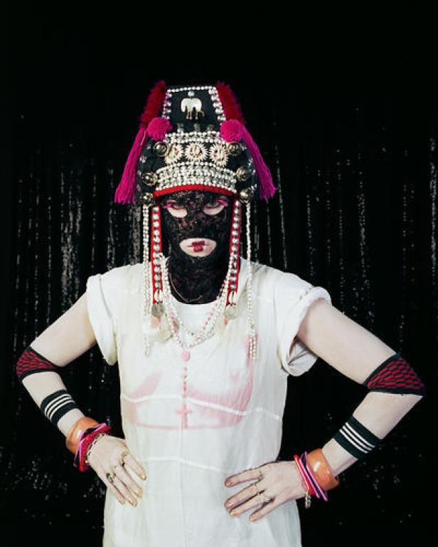 Eine Person mit aufwendigem Kopfschmuck und Spitzenmaske vor einem dunklen Hintergrund.