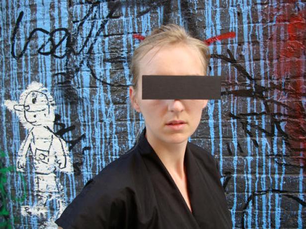 Eine Frau steht vor einer mit Graffiti bemalten Wand, ihre Augen sind mit einem schwarzen Balken bedeckt.