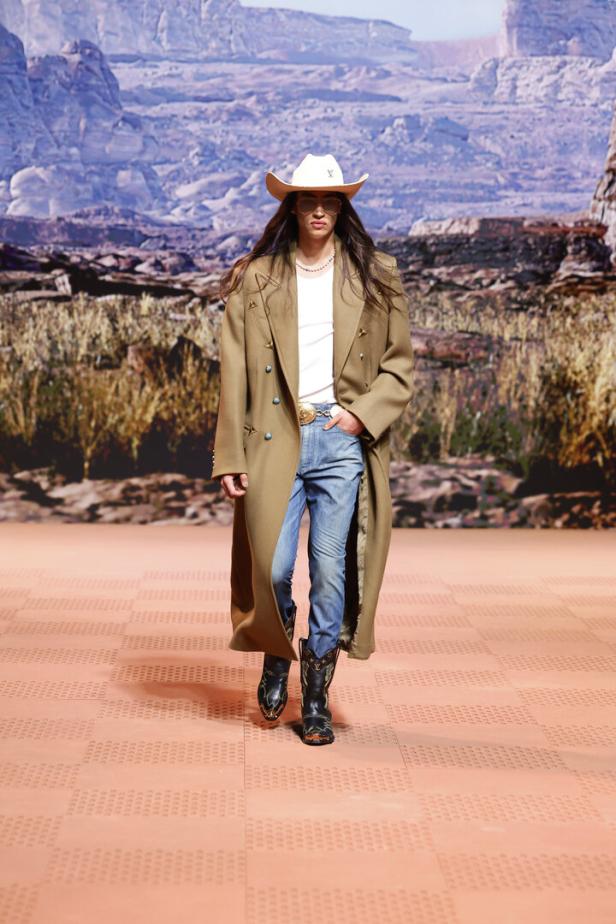 Ein Model in Cowboyhut, Mantel und Stiefeln läuft auf einem Laufsteg vor einer Wüstenlandschaft.