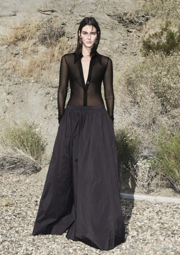 Eine Frau in einem transparenten schwarzen Hemd und einem langen, dunklen Rock posiert vor einer felsigen Landschaft.