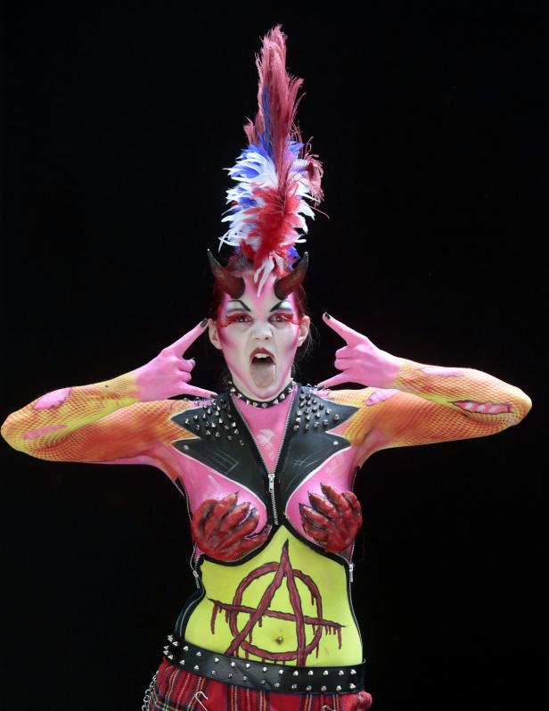 Eine Frau mit Körperbemalung im Punk-Stil mit Hörnern und Irokesenschnitt posiert vor schwarzem Hintergrund.