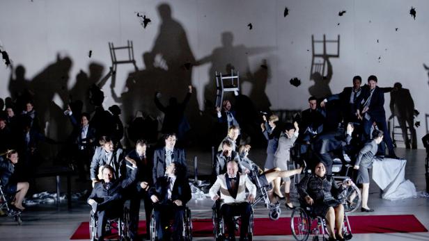 Eine Theaterszene mit Schauspielern in Anzügen, einige im Rollstuhl, vor einer Wand mit Schatten und Löchern.