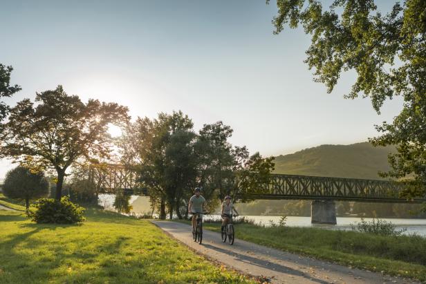 Ein Paar fährt mit Fahrrädern auf dem Donauradweg an einer Brücke vorbei.