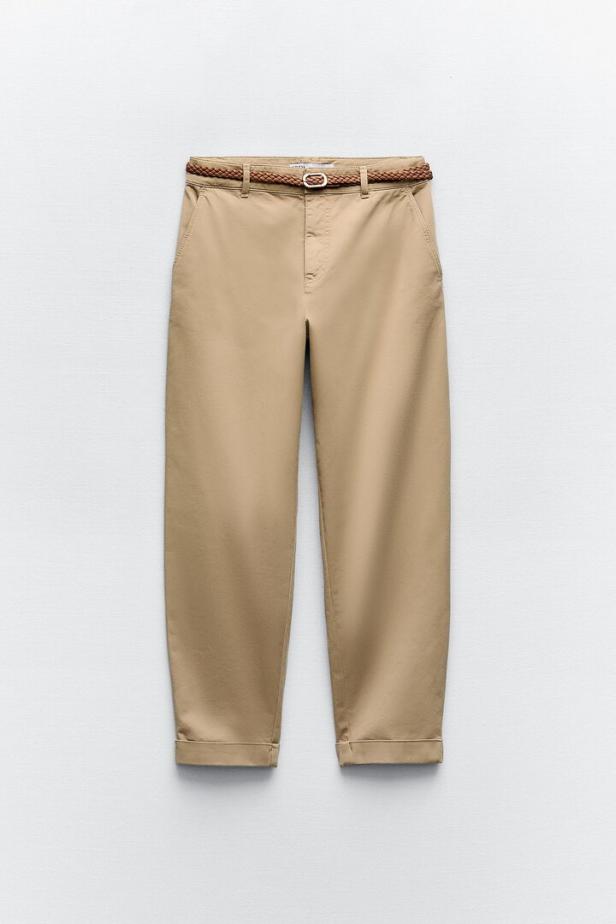 Eine beige Hose mit einem geflochtenen Gürtel vor einem neutralen Hintergrund.
