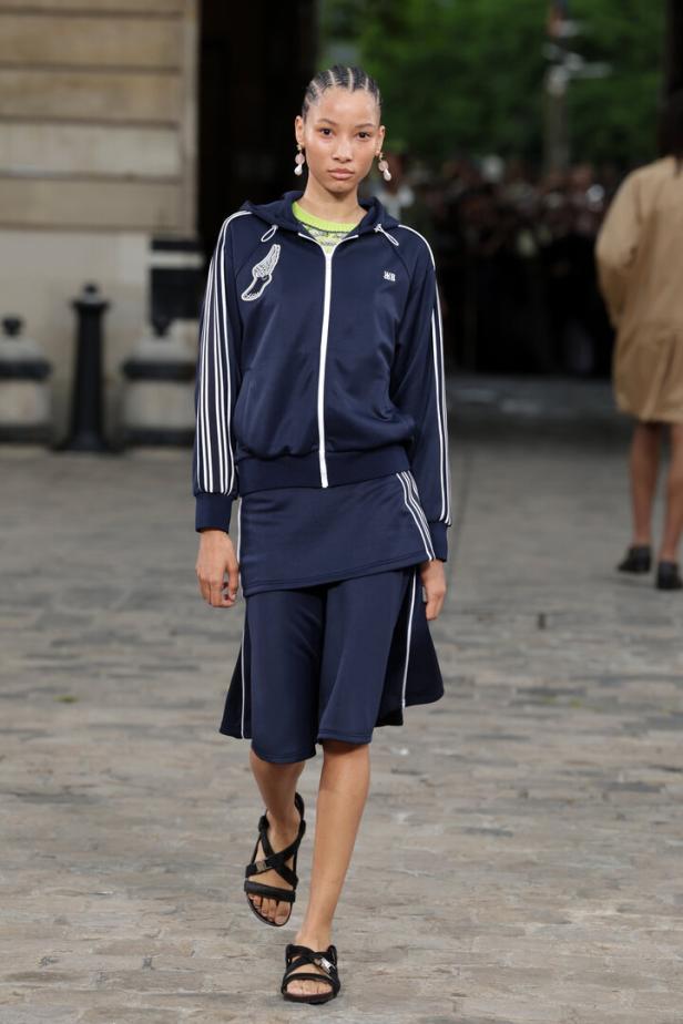 Eine Frau präsentiert ein marineblaues Trainingsoutfit mit weißen Streifen und Sandalen.