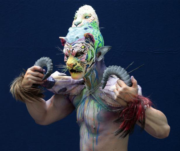 Ein Mann mit aufwendiger Körperbemalung und einem Kopfschmuck aus Tiermasken posiert vor einem blauen Hintergrund.