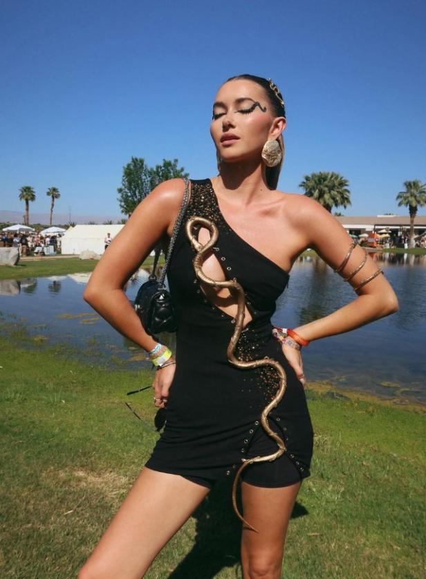 Eine Frau in einem schwarzen Kleid mit einer goldenen Schlange posiert vor einem See.