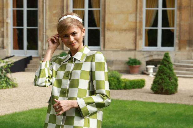 Zendaya trägt einen grün-weiß karierten Mantel und ein Haarband vor einem Gebäude.