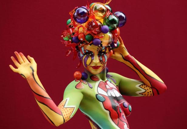 Eine Frau mit aufwendiger Körperbemalung und einem Kopfschmuck aus bunten Kugeln vor rotem Hintergrund.