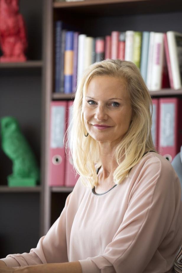 Eine blonde Frau sitzt vor einem Bücherregal mit bunten Büchern und Ordnern.