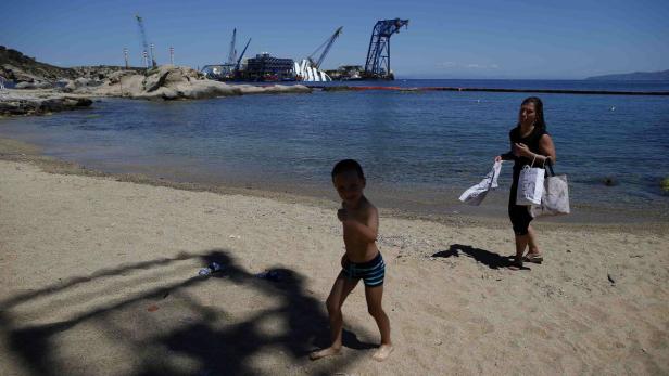 Ein Junge und eine Frau sind an einem Strand; im Hintergrund ist das Wrack der Costa Concordia zu sehen.