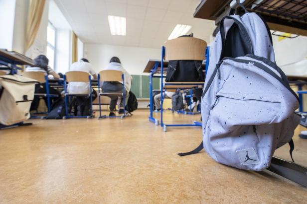Ein hellgrauer Rucksack steht im Vordergrund eines Klassenzimmers.