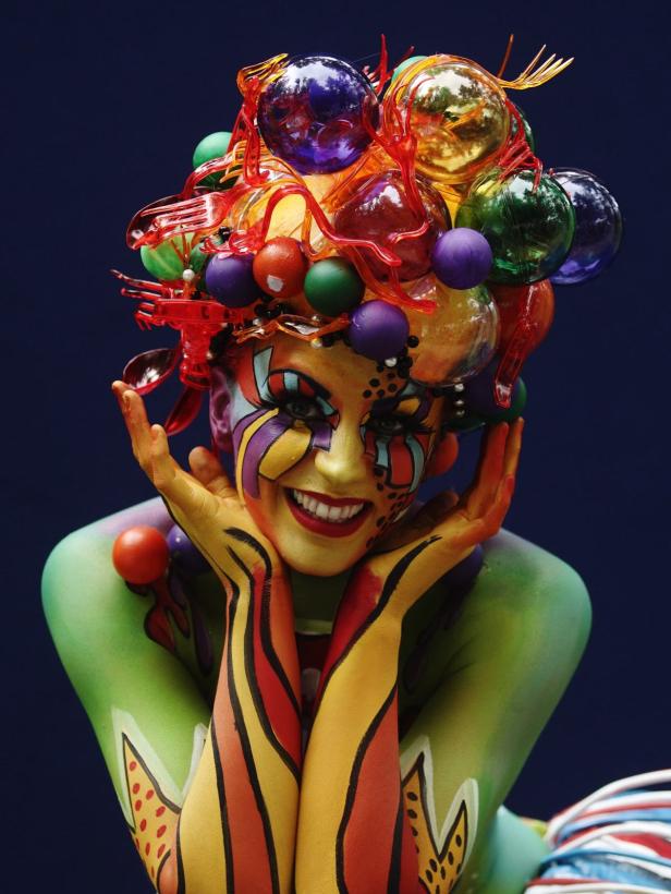 Eine Frau mit farbenfroher Körperbemalung und einem extravaganten Kopfschmuck lächelt.