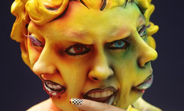 Ein Gesicht mit mehreren Gesichtern, bemalt in Gelb-, Grün- und Rottönen, mit unterschiedlichen Gesichtsausdrücken.