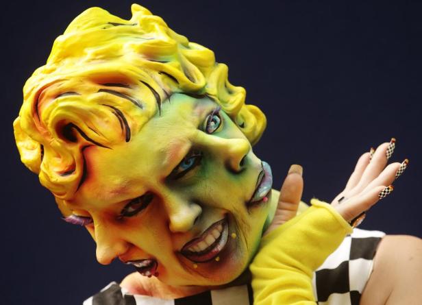 Eine Person mit aufwendigem, gelb-grünem Make-up und zwei Gesichtern posiert vor dunklem Hintergrund.