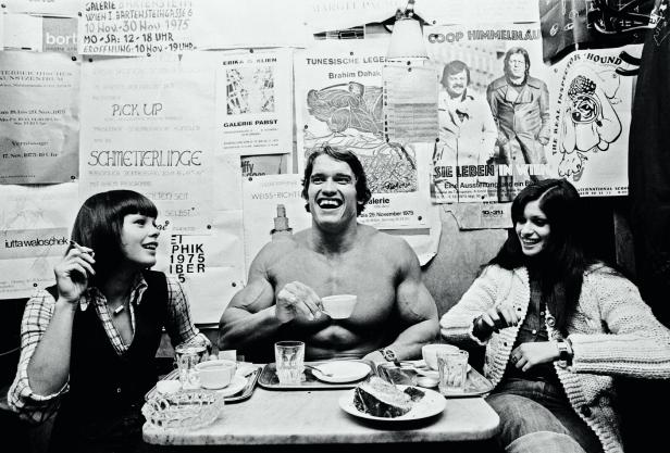 Arnold Schwarzenegger sitzt mit zwei Frauen in einem Café und trinkt Kaffee.