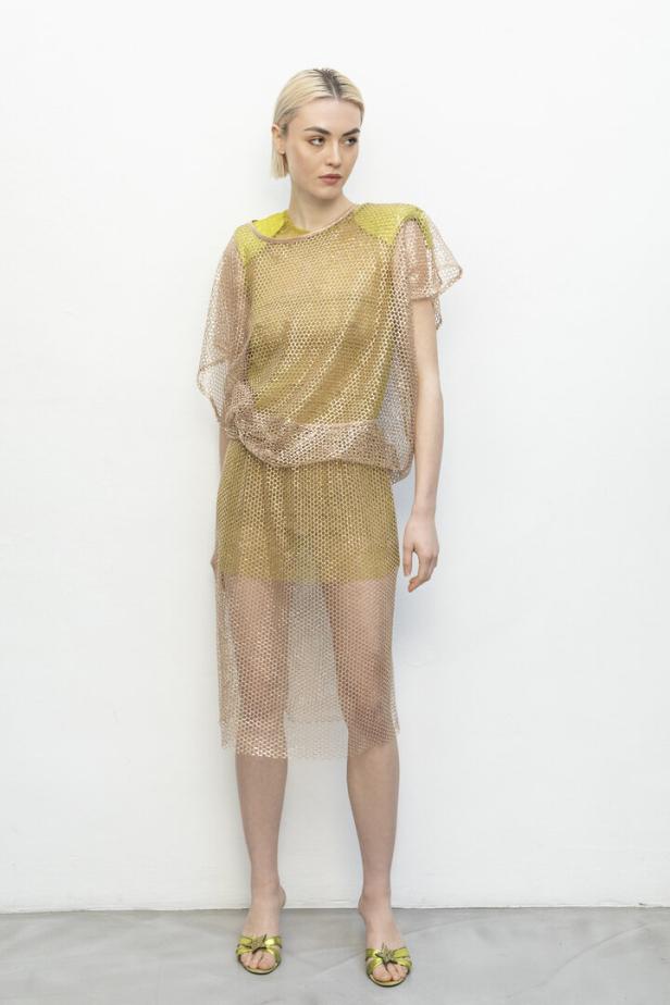 Eine Frau mit blonden Haaren trägt ein goldenes Netzoberteil und einen Rock mit grünen Schuhen.