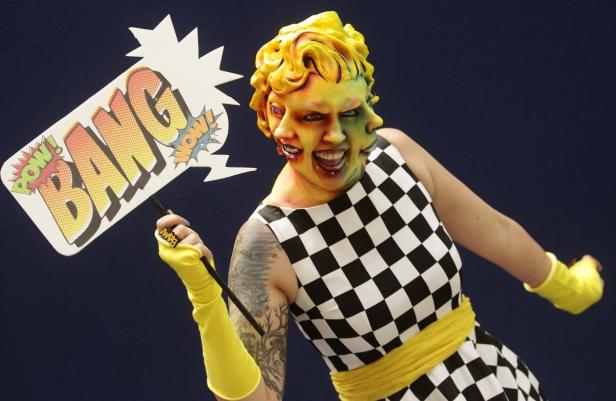 Eine Person mit aufwendigem Make-up und einem Schild mit der Aufschrift „BANG“.