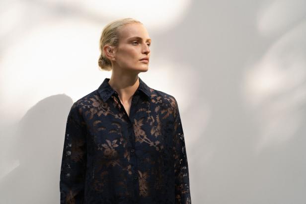 Eine Frau mit blonden Haaren trägt eine dunkelblaue, transparente Bluse mit Blumenmuster.