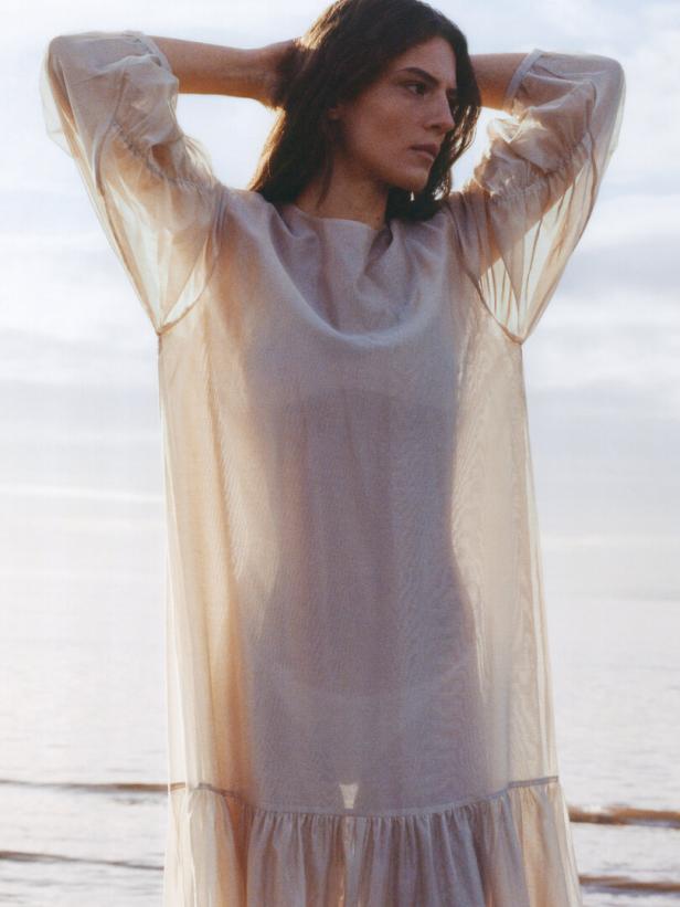 Eine Frau in einem transparenten Kleid steht am Strand und hält ihre Hände hinter dem Kopf.