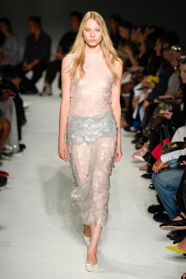 Ein Model präsentiert ein transparentes, helles Kleid mit Pailletten auf dem Laufsteg.
