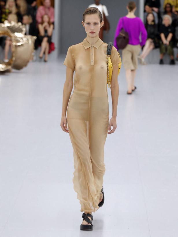 Ein Model präsentiert ein transparentes, beigefarbenes Kleid auf einem Laufsteg.