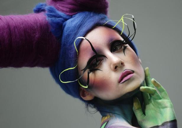 Eine Person mit aufwendigem Make-up, blauen Haaren und grünen Händen posiert.