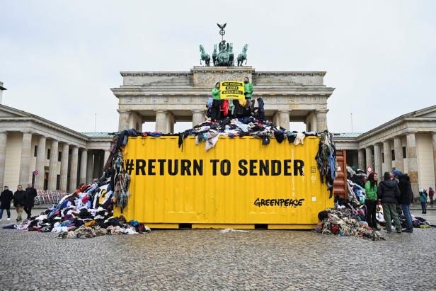 Vor dem Brandenburger Tor steht ein gelber Container mit der Aufschrift „#RETURNTOSENDER“, überfüllt mit Altkleidern.