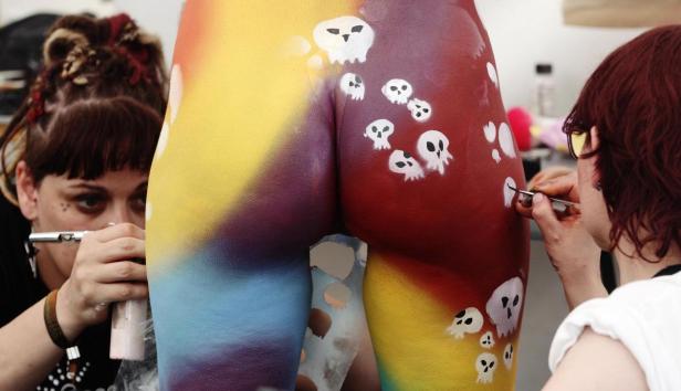 Zwei Künstlerinnen bemalen den Körper eines Models mit Regenbogenfarben und Totenköpfen.