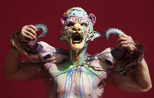 Eine Person mit Tiermaske, Körperbemalung und Schädeln mit Hörnern vor rotem Hintergrund.