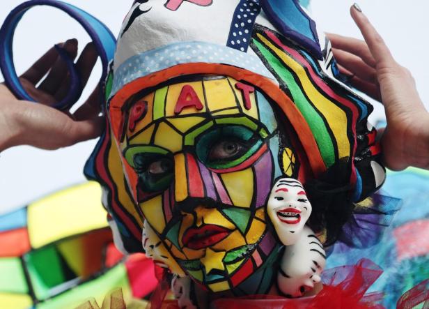 Eine Person mit aufwendiger, farbenfroher Gesichtsbemalung und kleinen Masken im Gesicht.