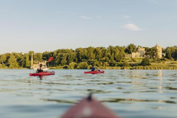 Zwei Personen paddeln in roten Kajaks auf einem See; im Hintergrund befindet sich ein Schloss.