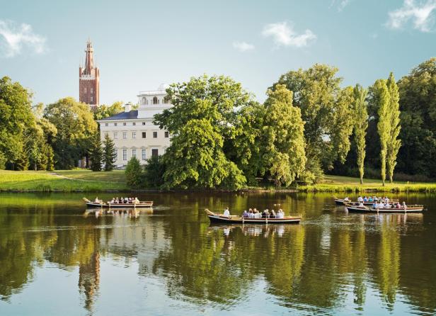 Anwesen inmitten eines grünen Parks mit einem See davor, auf dem Ruderboote mit Menschen schwimmen.