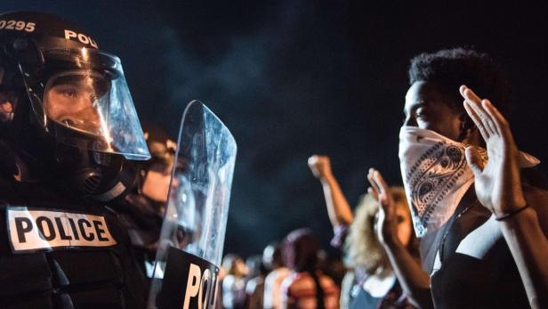 Ein Polizist in Schutzausrüstung steht einer Person mit erhobenen Händen gegenüber.