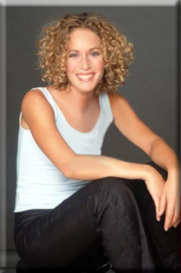 Eine lächelnde Frau mit blonden Locken sitzt in einem hellblauen Top und dunkler Hose.