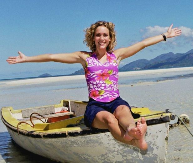 Eine Frau sitzt barfuß in einem Boot am Strand und streckt die Arme aus.