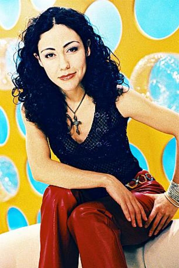 Eine Frau mit dunklen Locken, roter Hose und schwarzem Oberteil sitzt vor einem gelb-blauen Hintergrund.