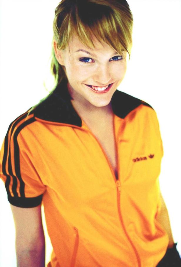 Eine lächelnde Frau in einer orangefarbenen Adidas-Trainingsjacke.