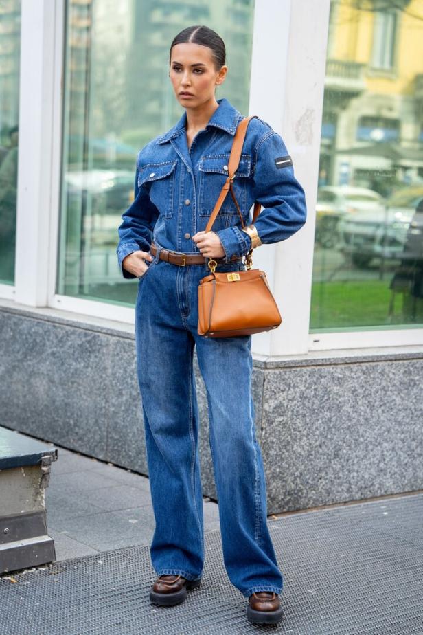 Eine Frau in einem Jeans-Overall mit brauner Handtasche und braunen Schuhen steht vor einem Gebäude.