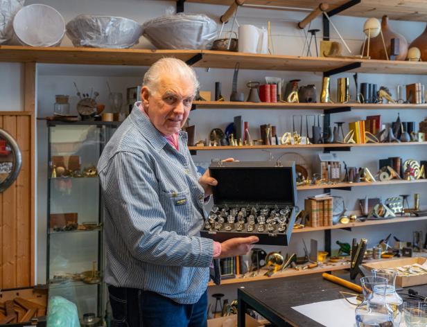 Carl Auböck IV mit gestreifter Jacke und Schachbrett in der Hand steht vor einem Regal voll mit Designobjekten des Unternehmens.