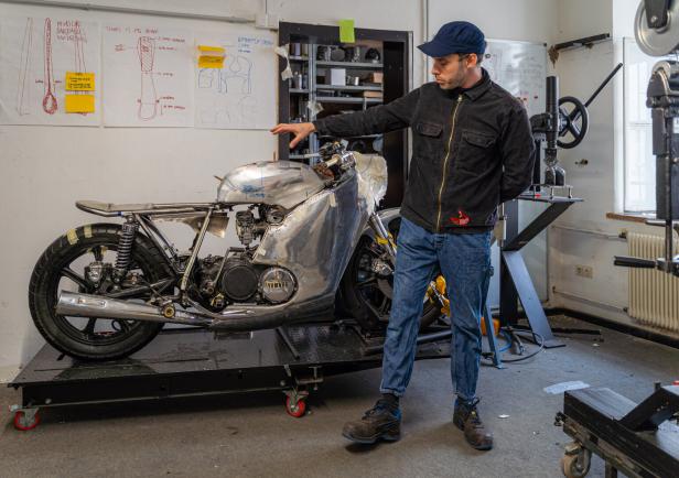 Ein Mann mit Kappe zeigt auf ein Motorrad, dessen Karosserie er selbst zusammengebaut hat.