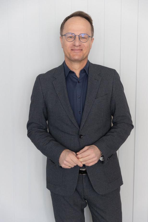 Ein Mann mit Brille und Anzug steht vor einer weißen Wand.